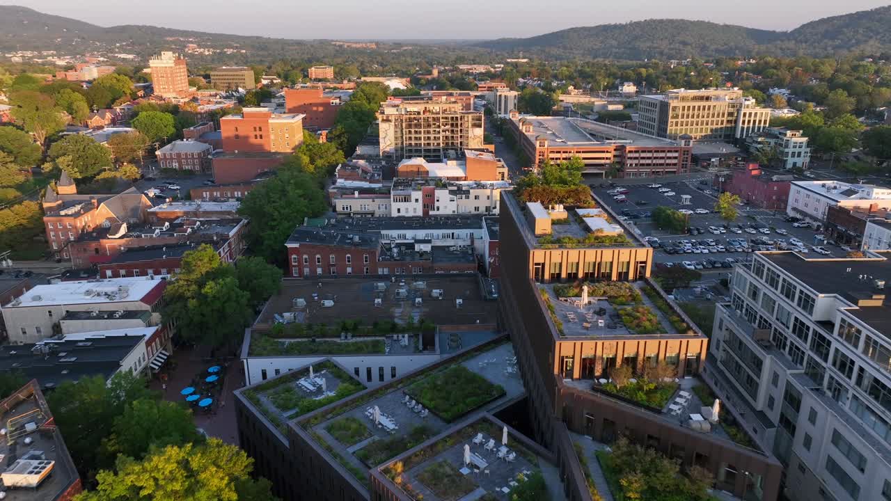 Downtown streets with historic buildings surrounded by trees and hills. Parking lots and rooftops fill urban landscape. Aerial view of Charlottesville, Virginia.