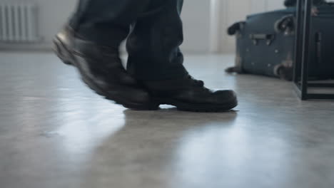 low angle view of person wearing black boots and dark trousers walking toward seat with travel bag nearby on smooth floor in bright room with subtle reflection and minimal background