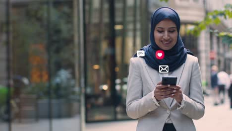 smiling muslim businesswoman standing outside city offices looking at mobile phone with motion graphics emojis showing multiple networking messaging and social media notifications