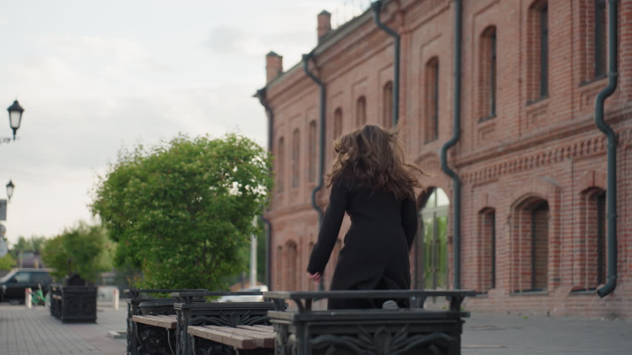 Woman in long coat stands near bench holding neck thoughtfully while handbag rests beside under soft daylight creating emotional urban scene with depth of field and architectural texture