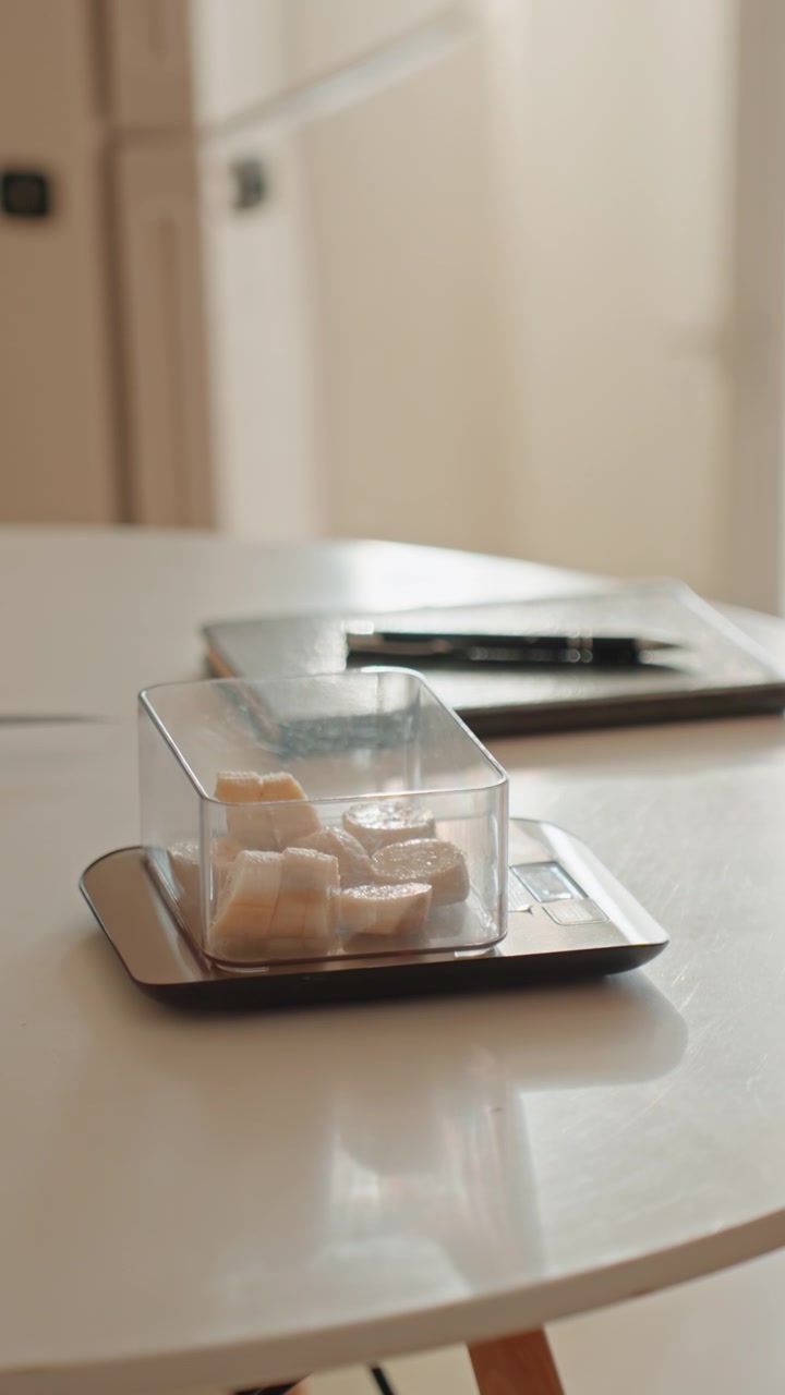 Home kitchen weighing banana slices for fitness, womans hands place transparent container on digital scale for precise portioning, soft natural light over white table, delicate bracelet and smartwatch