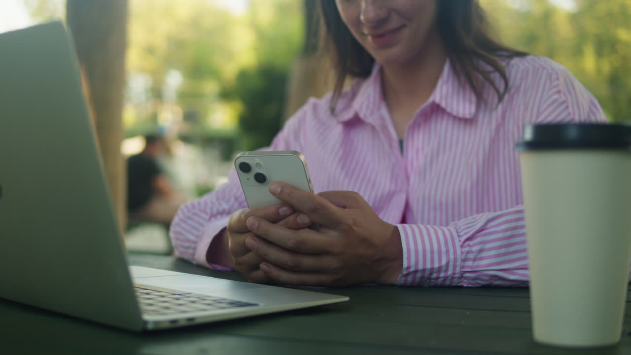 Woman is chatting with friends with phone in the park. Student girl sitting outside drinking coffee and communicating wit smartphone.
