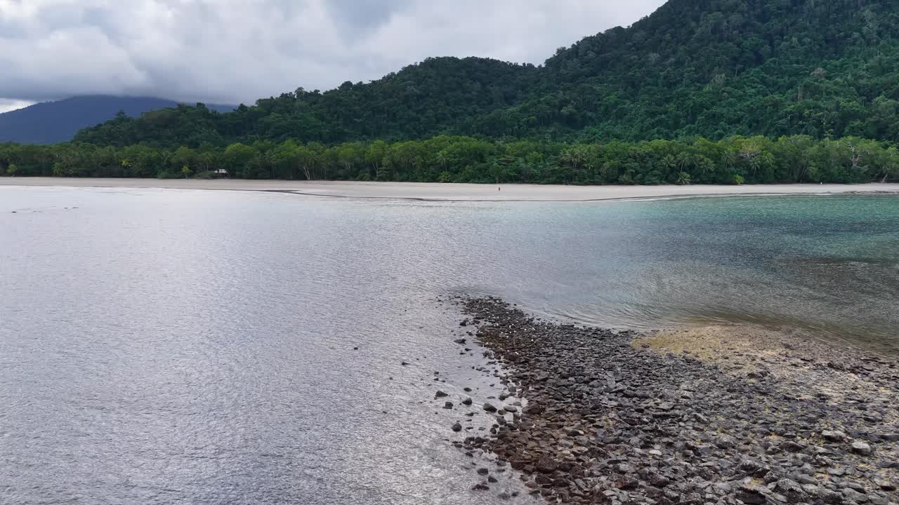 Aerial drone footage glides above a rocky shoreline and calm coastal waters, revealing lush rainforest hills under overcast skies in Port Douglas, Queensland