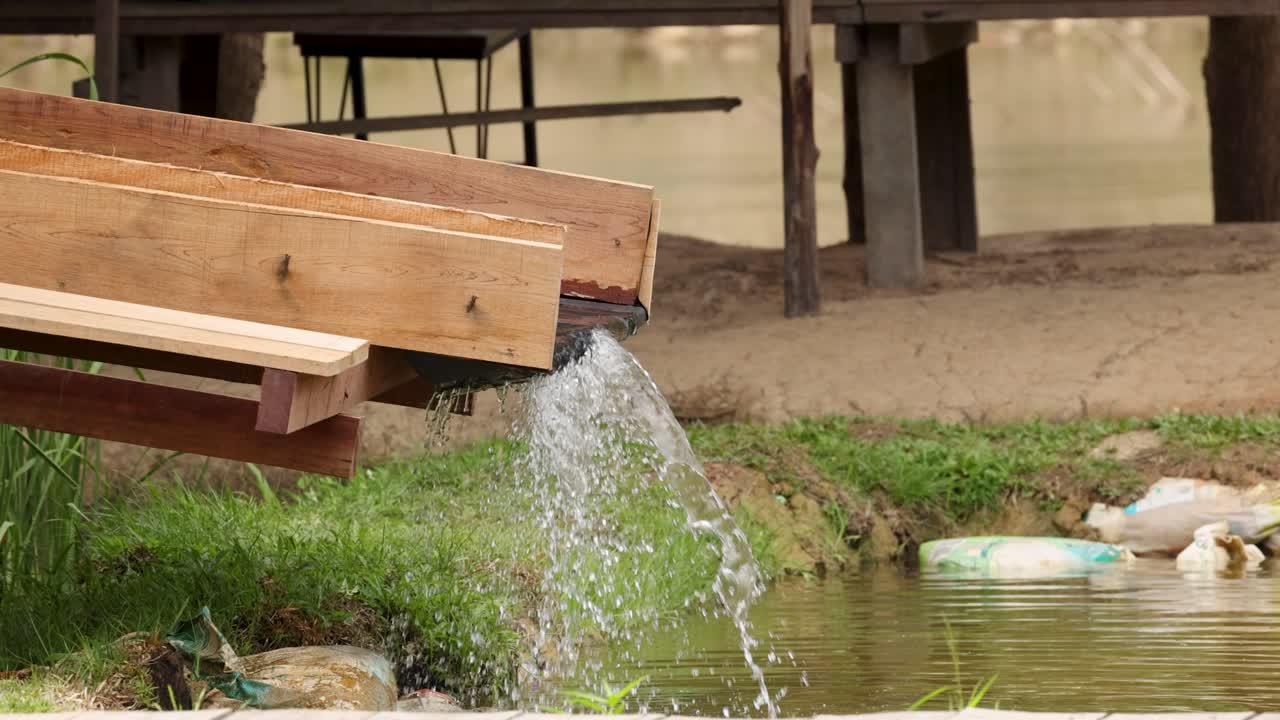 A wooden trough channels water into a pond, surrounded by grass and a rustic environment, under natural daylight