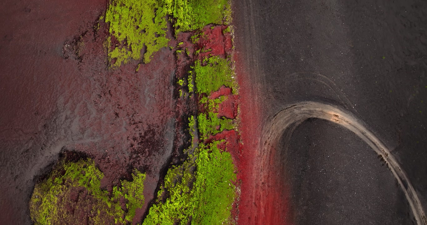 Top-down aerial view of a curved ash road cutting through vivid red volcanic earth and bright green moss, revealing striking natural color contrast and abstract geothermal patterns.