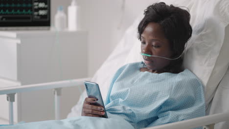 a young woman writes a message on her phone while lying in a hospital ward. an african girl is lying in a ward connected to ecg and oxygen devices in a mask and writes messages to relatives