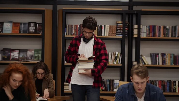 Slow motion footage of a guy in plaid shirt and headphones walking by row with pile of books and reading. Other students sitting, studying in college, university library. Front view