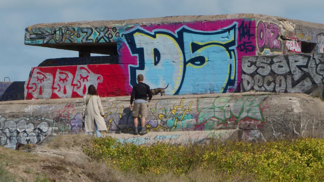 Two people examine a colorful graffiti-covered WWII bunker under bright daylight in Dunkirk