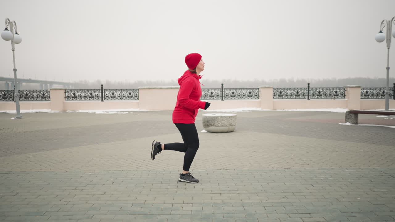 Side view of woman jogging outdoors in red hoodie and beanie on interlocked pavement during winter, scenic urban background with decorative railings, snow, and minimalist surroundings