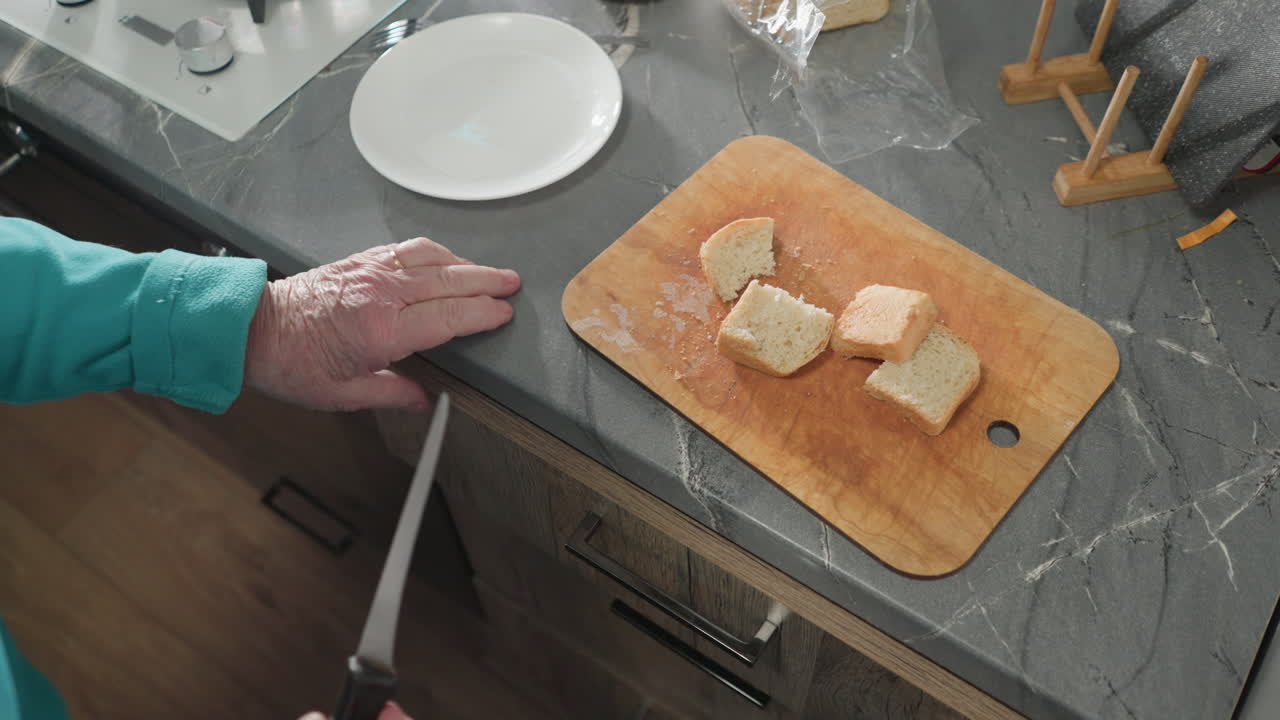 Close-up of person slicing soft bread on wooden cutting board in kitchen, kitchenware drawer open with utensils. Bread loaf wrapped in plastic beside cut pieces of bread