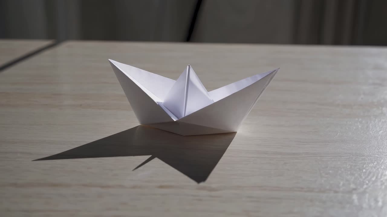 Delicate white origami boat resting motionless on wooden surface, casting subtle shadow shifting gently with changing light's angle, embodying minimalist paper craft design
