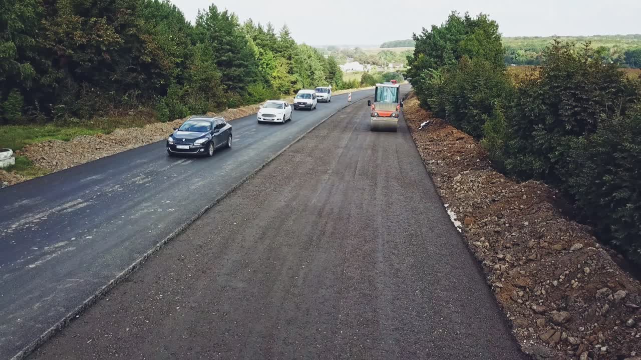 Vibratory roller compresses fresh asphalt on one side of the road on the background of the movement of cars on the other side of the road. Modern equipment for road construction works. Aerial view.