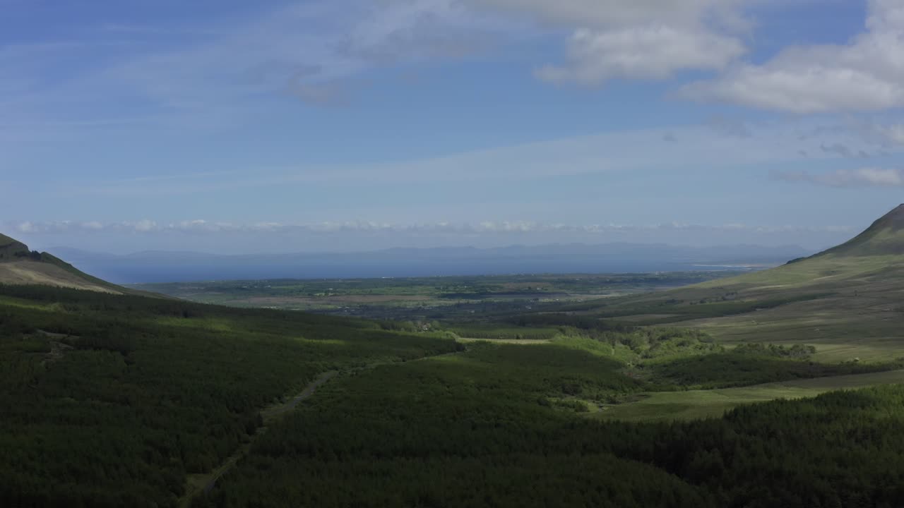 Gleniff Horseshoe valley, Sligo, Ireland, June 2021. Drone looking north with Donegal mountains on the horizon while slowly pulling backwards over rugged fields and woodlands.
