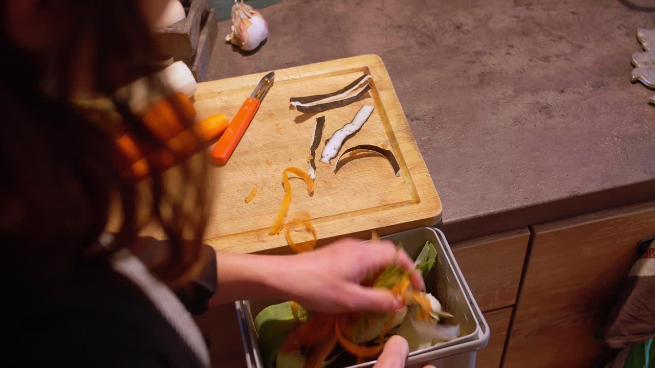 Eine Person sammelt Gemüsereste wie Karottenschalen in einem kleinen grauen Komposteimer auf einer hölzernen Küchenarbeitsplatte. Nahaufnahme.