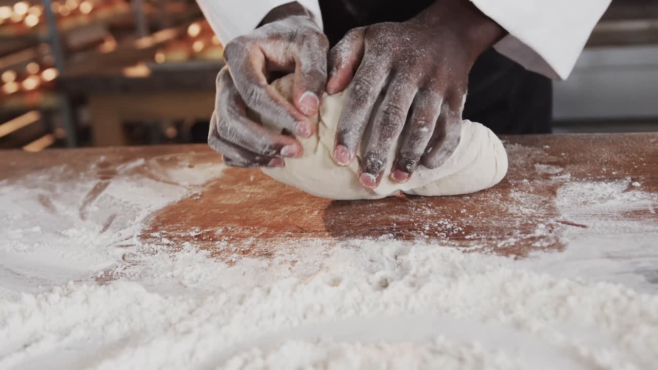 African american male baker working in bakery kitchen, kneading dough on counter in slow motion