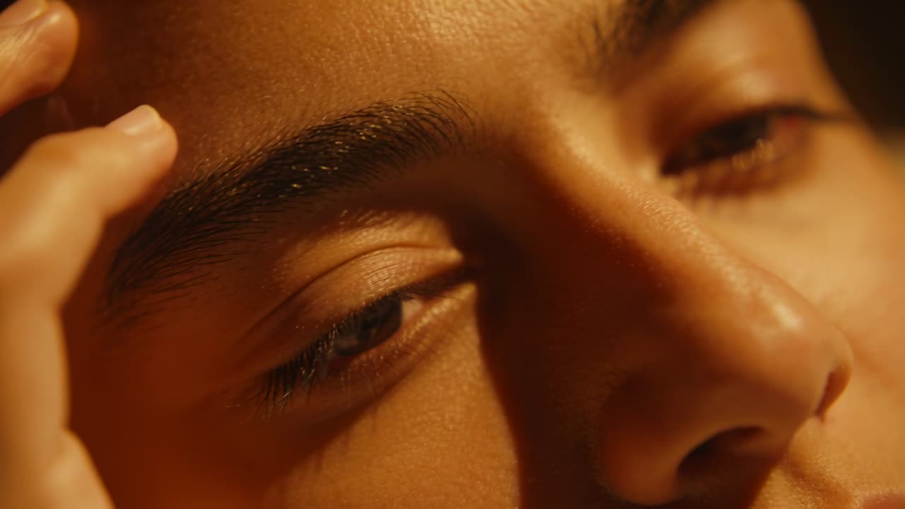 Hand entering left, touching female temple in dim room, eliciting eye opening under warm light