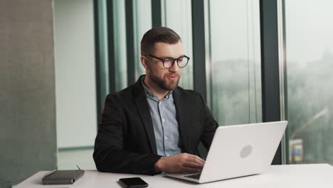 A handsome man in a business suit and glasses is talking and waving hello to a laptop