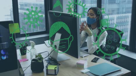 Animation-of-virus-icons-over-businesswoman-with-face-mask-using-computer-in-office