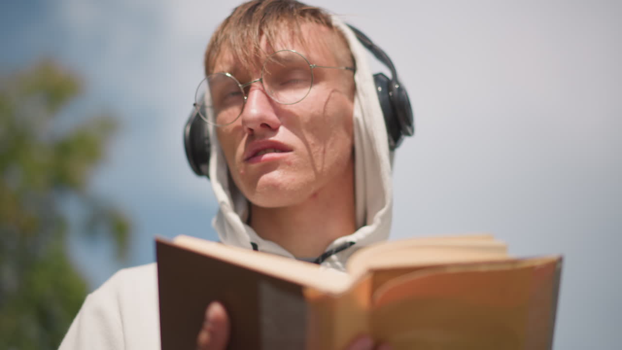 후드티를 입고 젖은 책을 읽고 있는 젊은 남성, 둥근 안경과 검은색 헤드폰을 착용, 얼굴과 페이지에 빗방울, 흐린 하늘의 은은한 빛, 야외 공원 배경, 집중한 표정과 내성적인 분위기