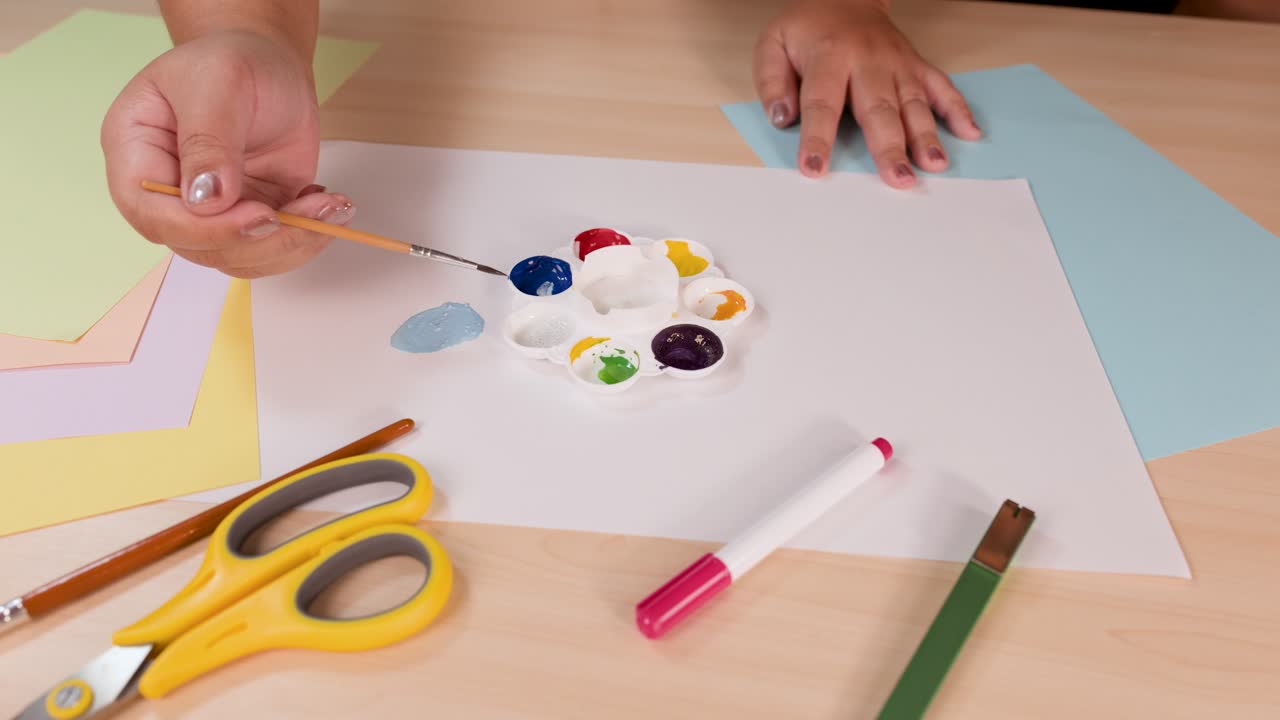 Person mixes watercolor paints with brush on palette, bright desk, overhead view, soft lighting