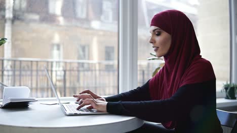 Ein-Junges-Attraktives-Muslimisches-Mädchen-Mit-Hijab,-Das-Ihren-Kopf-Bedeckt,-Tippt-Etwas-Auf-Ihrem-Laptop,-Während-Sie-In-Einem-Angeblichen-Co-Working-Bereich-Mit-Glasfenstern-Im-Hintergrund-Sitzt