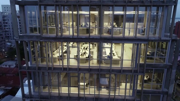 Drone sobe ao lado da fachada de um escritório moderno, revelando espaços de trabalho internos iluminados para conteúdo corporativo e imobiliário