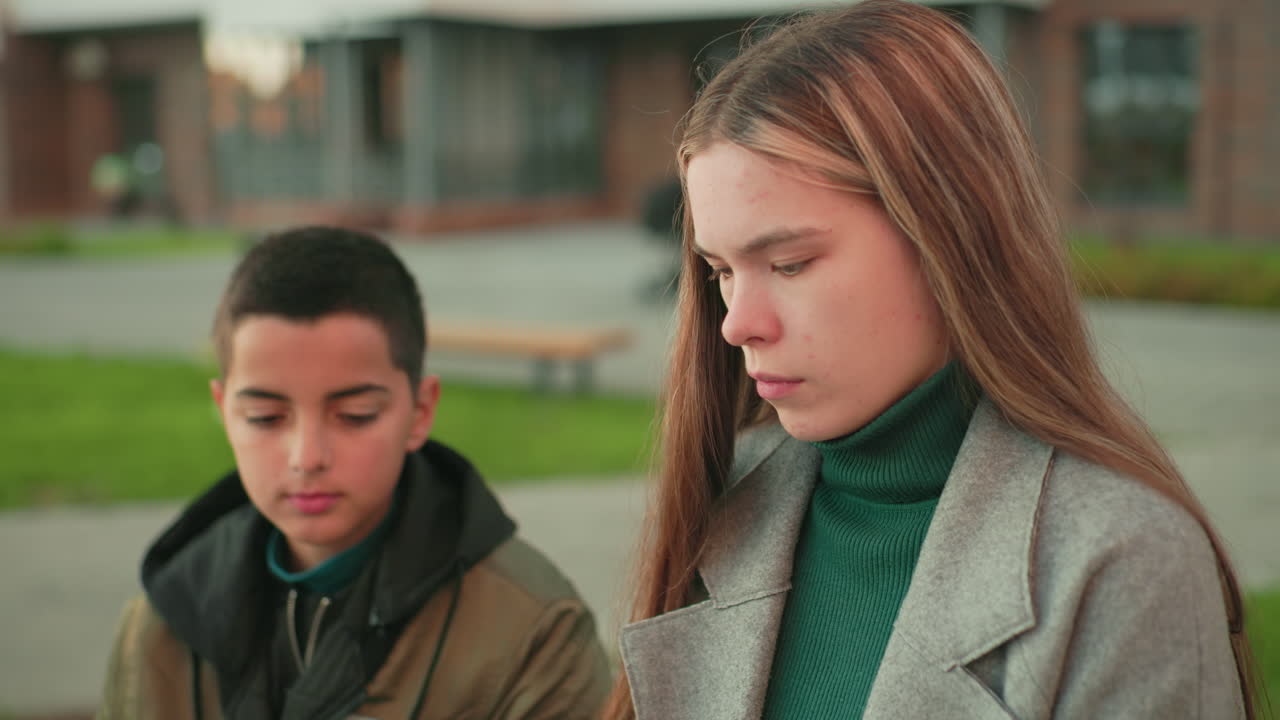 Boy attentively watches sister focusing on task while blurred passerby moves in background, both seated outdoors with calm expressions, capturing moment of learning