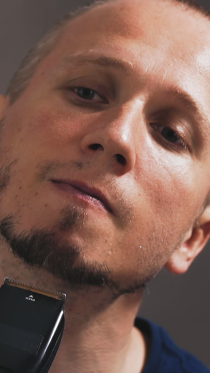 close up slow motion young unshaven guy looks at himself in mirror, is going to shave off his beard with an electric hair clipper, thinks long, winces, does not dare to start