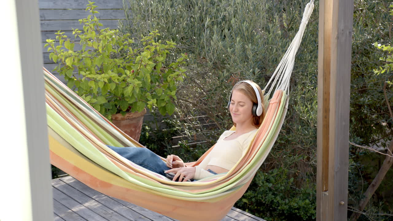 Relaxing in hammock, woman listening to music with headphones and using tablet