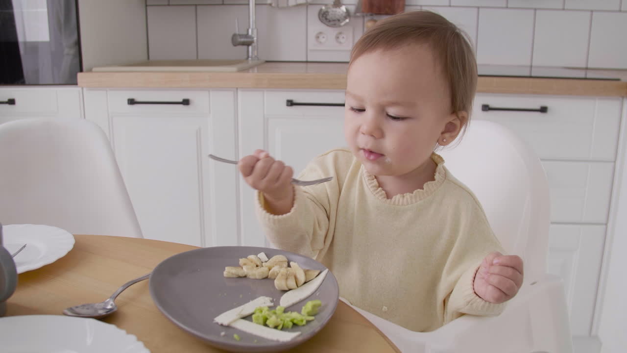 Sød lille pige sidder i højstol ved siden af køkkenbordet og spiser mad med gaffel og hænder.