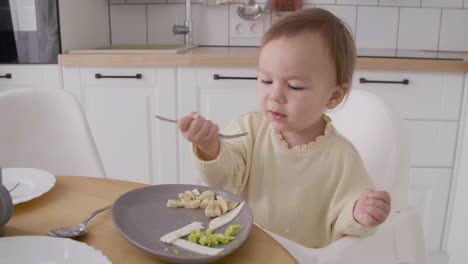 cute baby girl sitting in high chair next to the kitchen table eating food with fork and hands