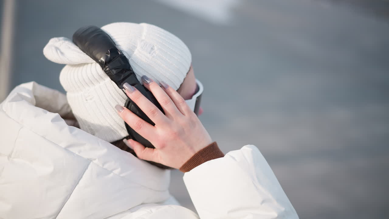 Young girl wearing white beanie and puffer coat grooving to music under bright winter sun while adjusting over ear headphones with long painted nails on snow covered urban plaza lively scene