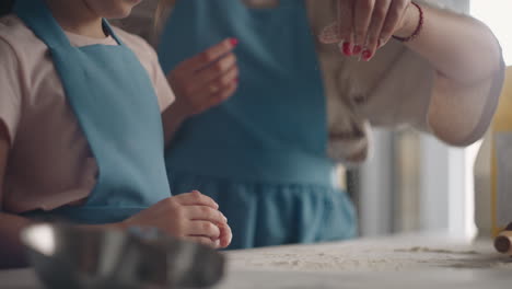 mother is teaching little daughter to make bread in home child is kneading dough on table