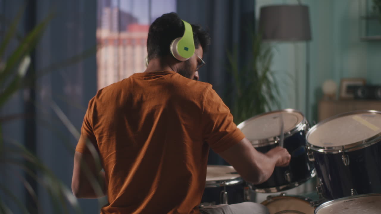 Indian Man Playing Drums in Evening Indian Drummer in Casual Clothes Listening to Music in Wireless Headphones and Playing Drums during Rehearsal in Living Room in Evening