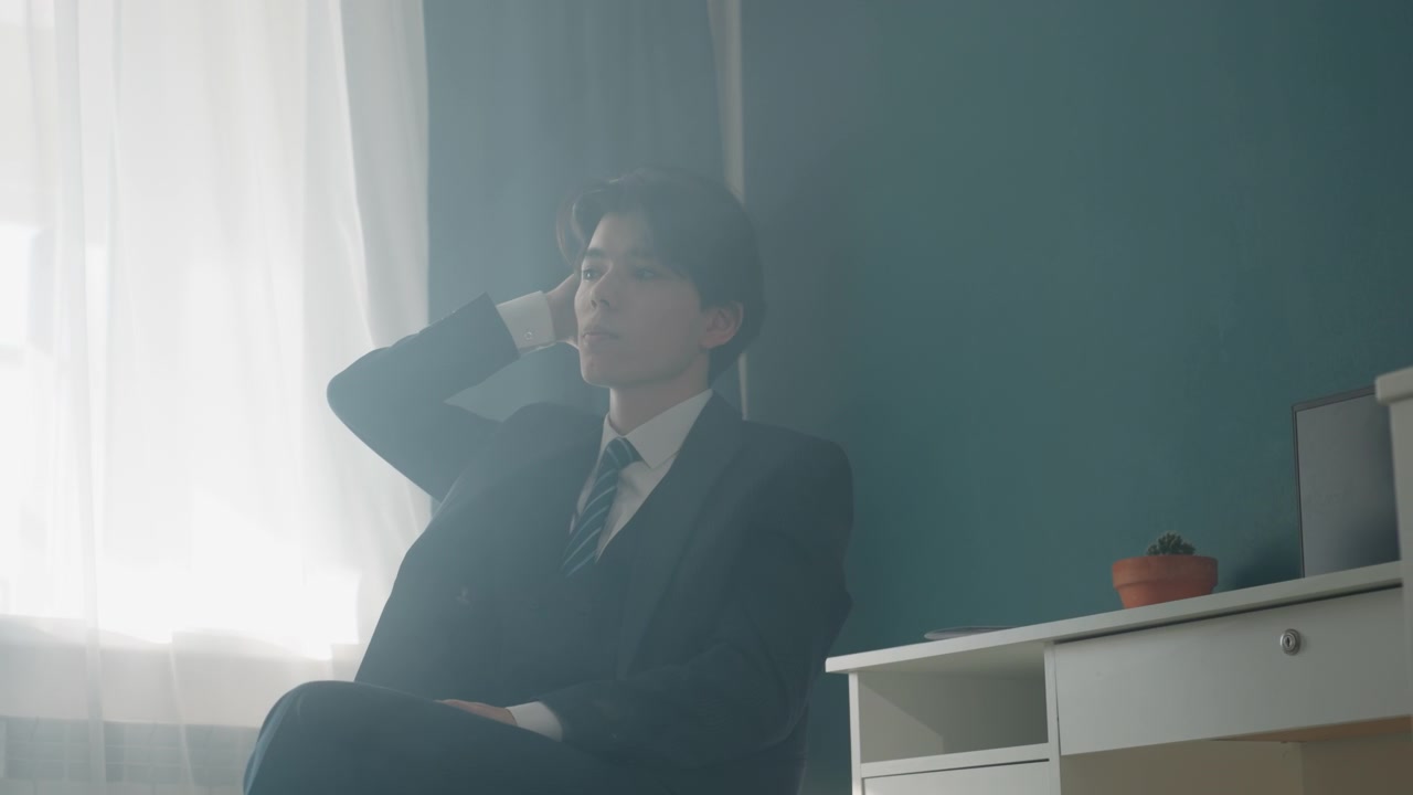 穿著西裝的商務人士坐在窗邊，在薄紗窗簾透入的柔和陽光下沉思，書桌上擺放著小盆栽，藍綠色牆壁，電影般的薄霧和輕柔的陰影，若有所思的姿態。