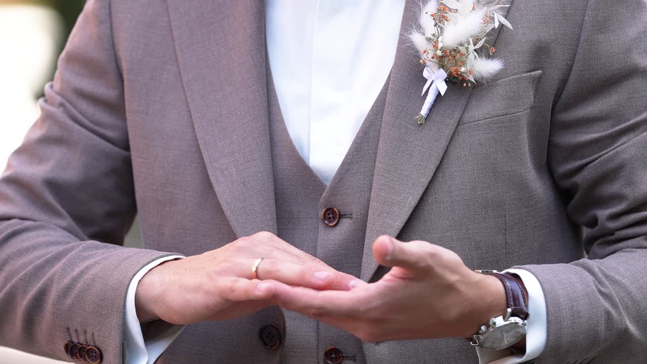 Close-up of groom's hands in stylish brown suit adjusting his jacket, showcasing a serene pre-wedding moment. Perfect lighting highlights the delicate boutonniere detail