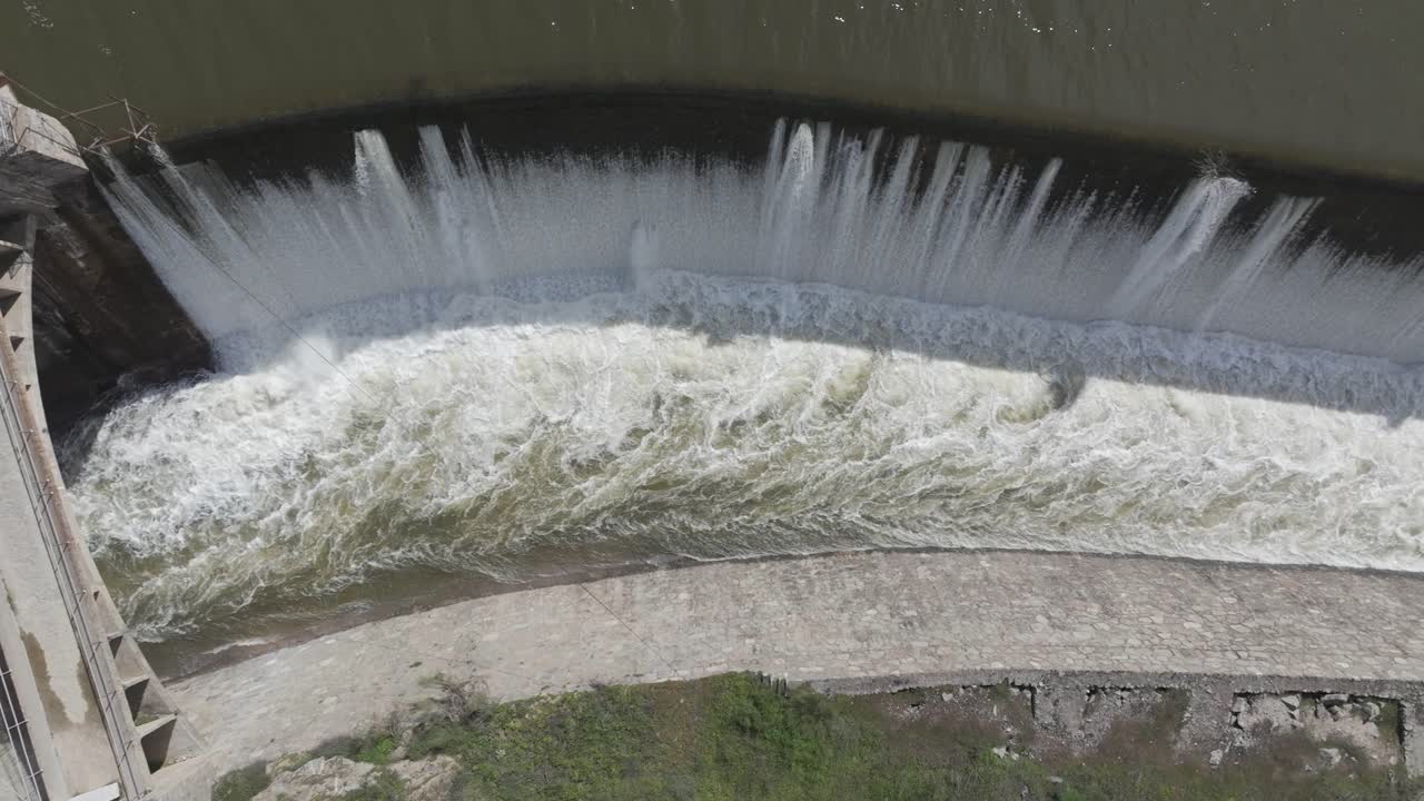 Slow-motion top-down drone footage of a dam's spillway. A bridge appears first, then water pours through the lateral chute forming a stunning waterfall into a wide canal. Truly impressive.
