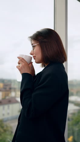 calm caucasian lady in black jacket standing at window with a cup in hands. happy woman having a break from work for coffee. vertical video.