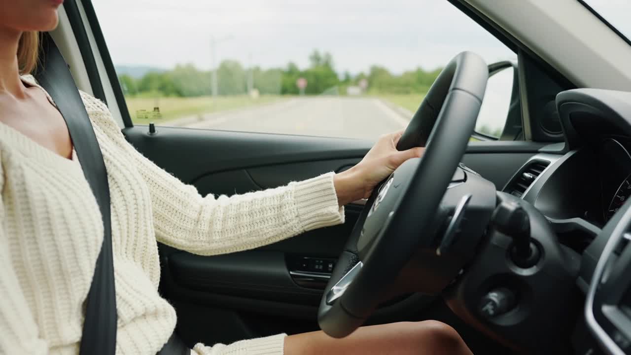 Una donna con un maglione color crema che gira la chiave, allaccia la cintura di sicurezza, impugna il volante e guida su una strada rurale.
