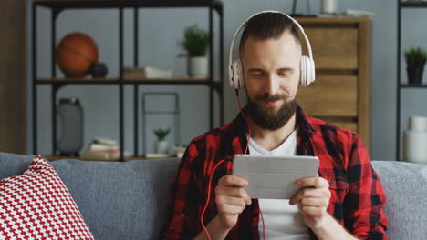 Close-Up-Of-The-Handsome-Hipster-Man-In-The-Big-White-Headphones-Watching-Something-Funny-On-The-Tablet-Computer,-Nodding-His-Head-And-Laughingon-The-Couch-In-The-Living-Room