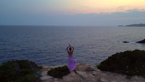woman meditating in yoga position on a cliff overlooking the sea at sunset in ibiza. overflight flyover amazing aerial view fligh drone