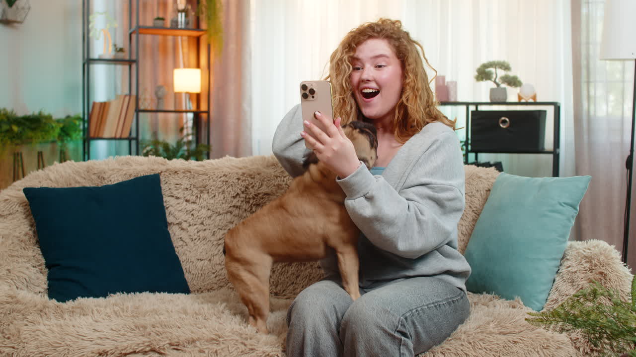 Young woman reacts happily to good news on smartphone with pug dog on home sofa smiling with emotion