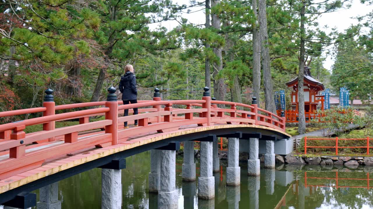 A cinematic scene capturing a blonde girl walking gracefully across a traditional bridge within the sacred grounds of Danjo Garan in Mount Koya, Koyasan, Japan.