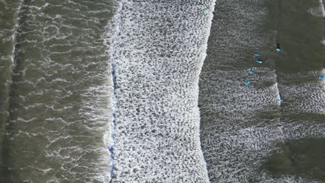 aerial view of foamy waves rolling onto a dark shore with scattered surfers waiting