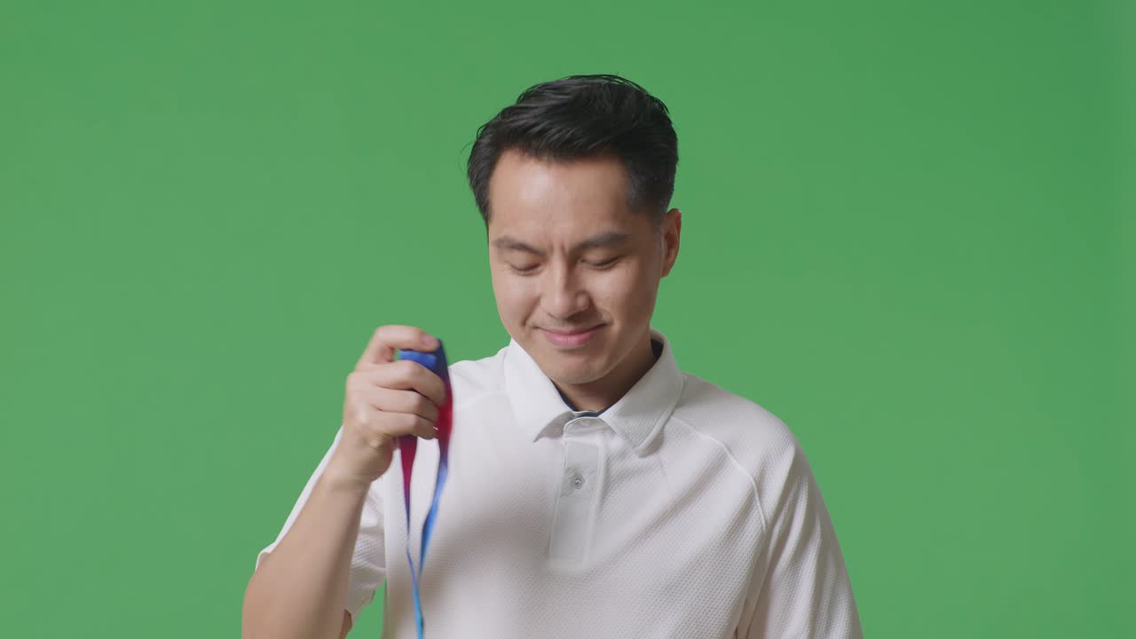 금메달을 입고 카메라를 향해 미소 짓는 아시아 남자가 스튜디오의 녹색 스크린 배경에서 첫 번째 우승자로 자부심을 느끼고 있습니다.
