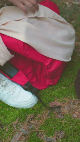 primer plano de damas en vaqueros y zapatillas de deporte recuperando mochilas del suelo del bosque, cerrándolas cuidadosamente antes de continuar su caminata, rodeadas de agujas de pino y musgo verde