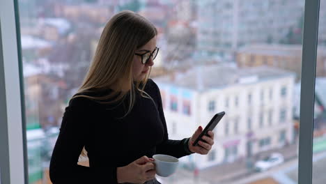 beautiful female with cup of coffee thoughtfully browsing smartphone while standing by the window