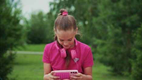 a girl in a pink shirt with matching headphones engrossed in a tablet, enjoying her time surrounded by lush greenery in a serene outdoor setting