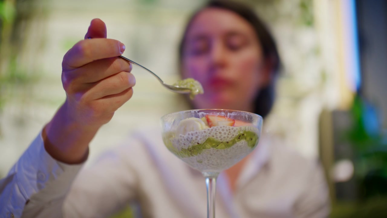Una donna tiene un cucchiaio di budino di chia davanti a un bicchiere da dessert riempito con budino di semi di chia bianchi, purea di kiwi verde, fragole a fette e banana.