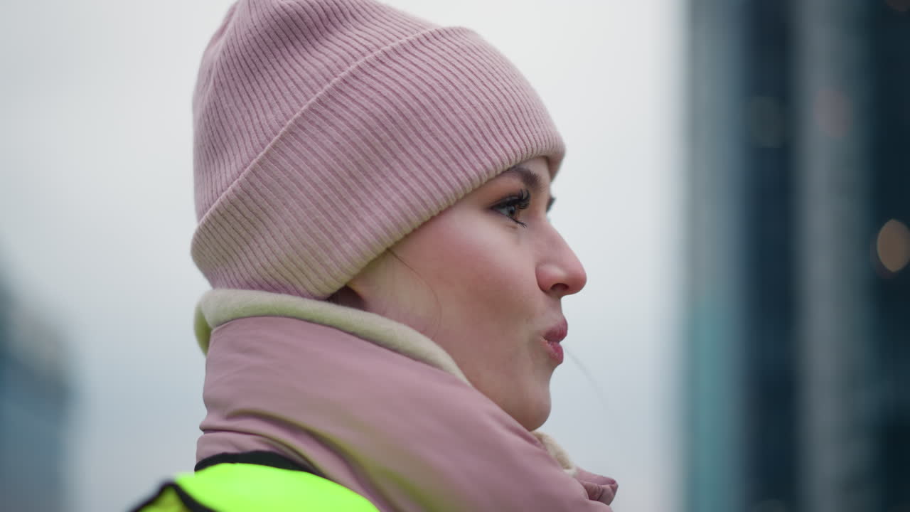 Young female worker in neon reflective vest and pink beanie smiling in profile during outdoor winter work, expressing warmth, friendliness while engaged in conversation in cold environment